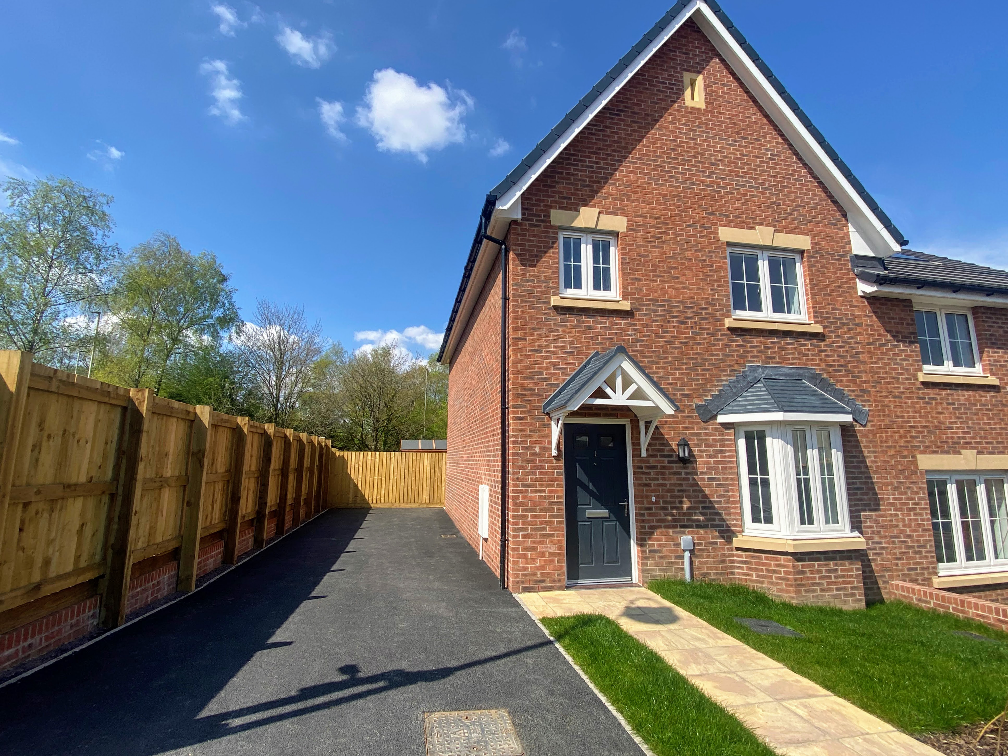 The exterior of one of our new homes in Hengoed, Caerphilly. The sky is blue, it has a large black tarmac driveway to the left. On the right is the house, with a black door and a bay window downstairs. There are two windows upstairs and a pointy roof. There is a cream patio out the front in a line from the door to the path, with grass in front of the window.